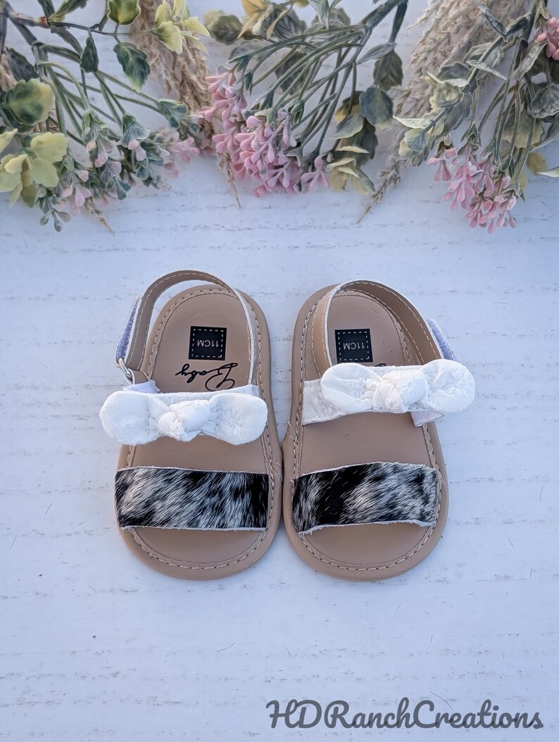 May include: A pair of baby sandals with a cowhide pattern on the toe strap. The sandals have a beige sole, white bow, and blue strap. The sandals are labeled "11cm" and "Body". The sandals are from "HDRanchCreations".