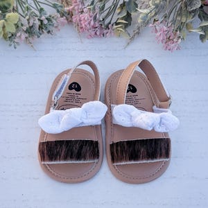 May include: A pair of baby sandals with a light brown base, a white bow, and a strip of brown fur across the toes. The sandals have a strap and are size 12. The sandals are on a white surface with floral decorations.