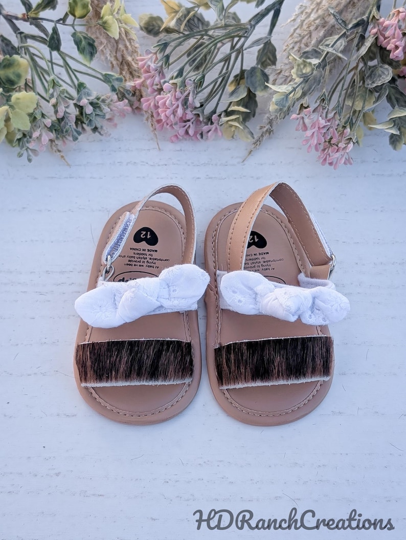 May include: A pair of baby sandals with a light brown base, a white bow, and a strip of brown fur across the toes. The sandals have a strap and are size 12. The sandals are on a white surface with floral decorations.