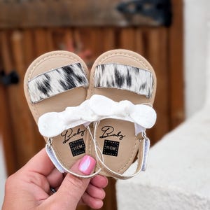 May include: A pair of baby sandals with a tan base, white straps, and a white bow. The sandals feature a black and white cowhide accent. The word "Baby" is printed on the insole, along with "11CM".