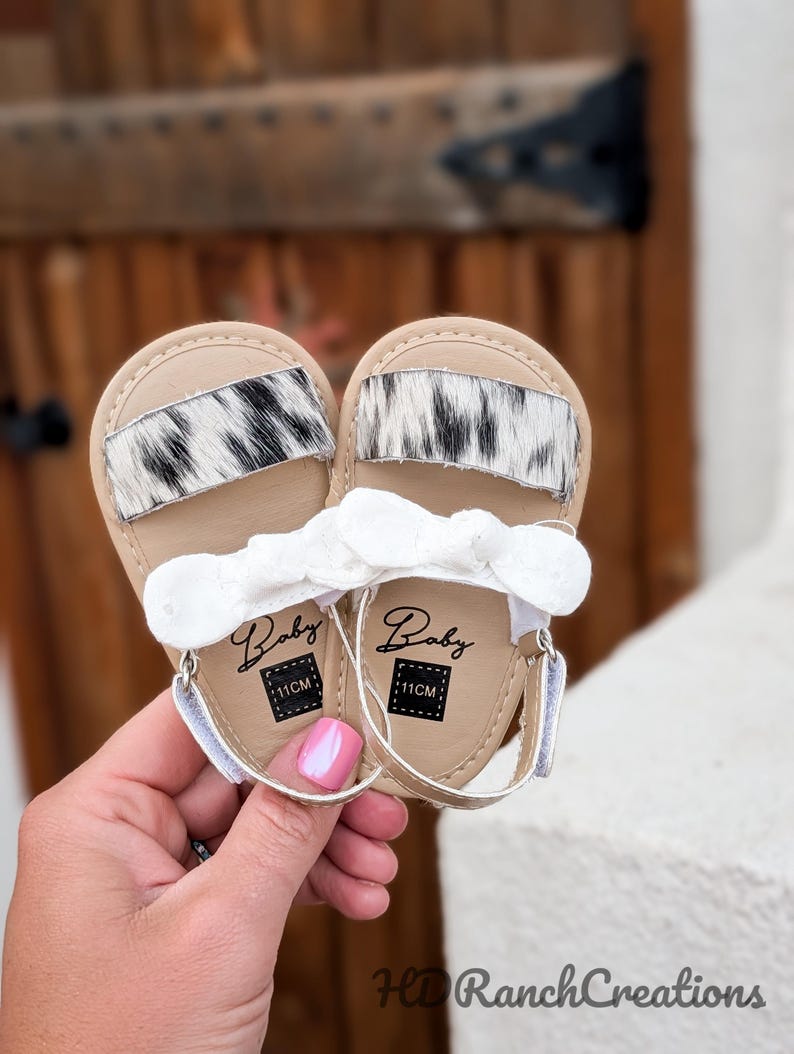May include: A pair of baby sandals with a tan base, white straps, and a white bow. The sandals feature a black and white cowhide accent. The word "Baby" is printed on the insole, along with "11CM".
