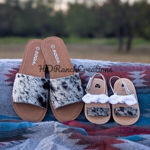 May include: A pair of adult and baby sandals. The adult sandals are black and white cowhide print with brown soles. The baby sandals have white ruffle accents. The sandals are displayed on a patterned blanket.