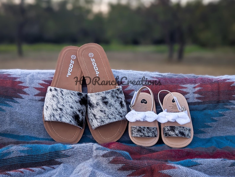 May include: A pair of adult and baby sandals. The adult sandals are black and white cowhide print with brown soles. The baby sandals have white ruffle accents. The sandals are displayed on a patterned blanket.