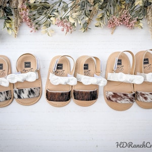 May include: Six pairs of baby sandals with cowhide accents and white ruffle details. The sandals have tan soles and straps, with a velcro closure. The cowhide patterns vary from black and white to brown and white. The sandals are arranged on a white surface.