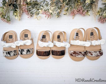 May include: Six pairs of baby sandals with cowhide accents and white ruffle details. The sandals have tan soles and straps, with a velcro closure. The cowhide patterns vary from black and white to brown and white. The sandals are arranged on a white surface.