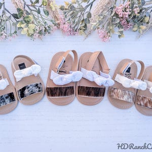 May include: Six pairs of baby sandals with tan straps and soles. The sandals have a cowhide pattern on the front and white bows. The sandals are arranged on a white surface with floral decorations.