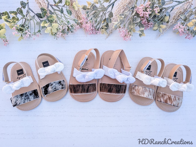 May include: Six pairs of baby sandals with tan straps and soles. The sandals have a cowhide pattern on the front and white bows. The sandals are arranged on a white surface with floral decorations.