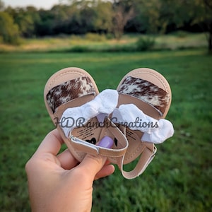 May include: Tan baby sandals with cowhide accents and white ruffled bows. The sandals have a strap closure and are held in a hand against a green background. The sandals are a baby shoe.