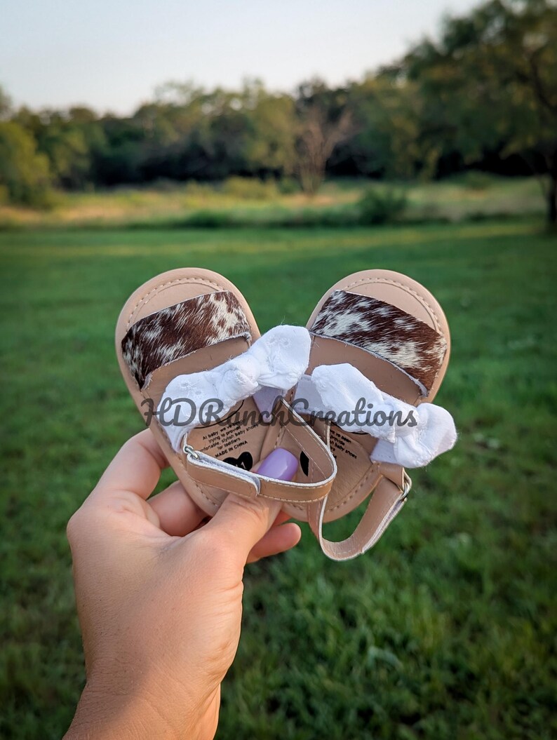 May include: Tan baby sandals with cowhide accents and white ruffled bows. The sandals have a strap closure and are held in a hand against a green background. The sandals are a baby shoe.