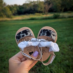 May include: Baby sandals with a cowhide print on the straps. The sandals have a light brown sole, white straps, and a silver buckle. The text "HDRanchCreations" is visible on the sandals.