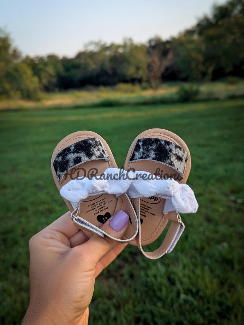 May include: Baby sandals with a cowhide print on the straps. The sandals have a light brown sole, white straps, and a silver buckle. The text "HDRanchCreations" is visible on the sandals.