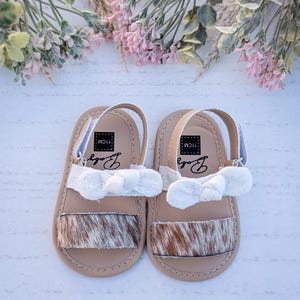 May include: A pair of baby sandals with a tan base, cowhide-style toe strap, and white bow accent. The sandals have a white strap and the word "Baby" printed on the insole. The sandals are 11cm long.