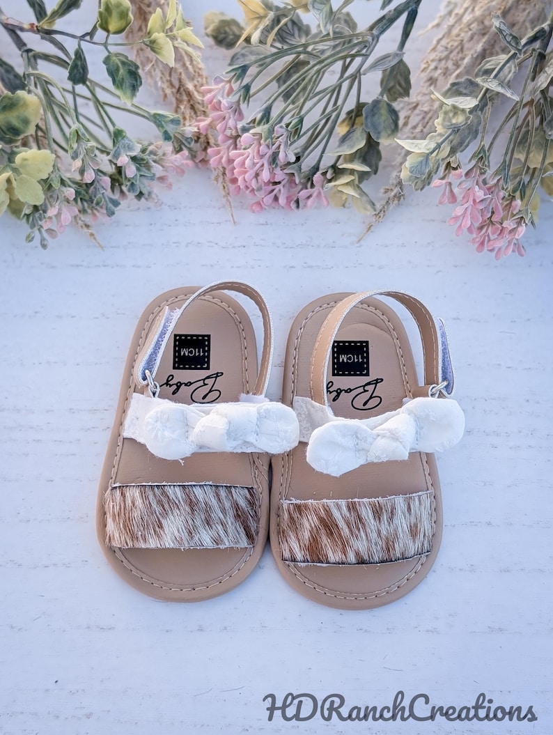 May include: A pair of baby sandals with a tan base, cowhide-style toe strap, and white bow accent. The sandals have a white strap and the word "Baby" printed on the insole. The sandals are 11cm long.