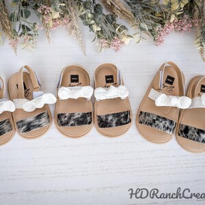 May include: Six pairs of baby sandals with a tan base, white straps, and a black and white cowhide pattern. Each sandal has a white bow. The sandals are arranged on a white surface with floral accents.