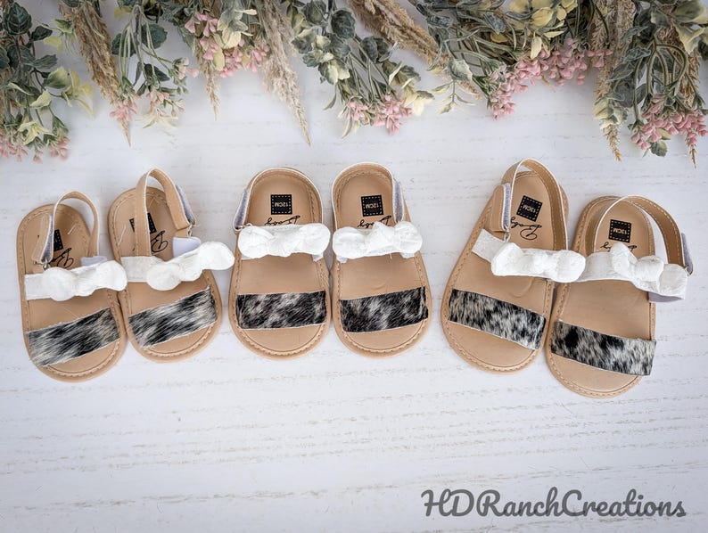May include: Six pairs of baby sandals with a tan base, white straps, and a black and white cowhide pattern. Each sandal has a white bow. The sandals are arranged on a white surface with floral accents.