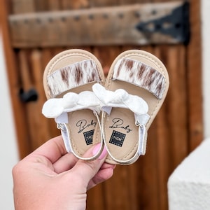 May include: A pair of baby sandals with a tan leather sole and a white strap. The sandals feature a brown and white cowhide design on the top strap and a white bow detail. The soles are marked "Baby" and "11CM".