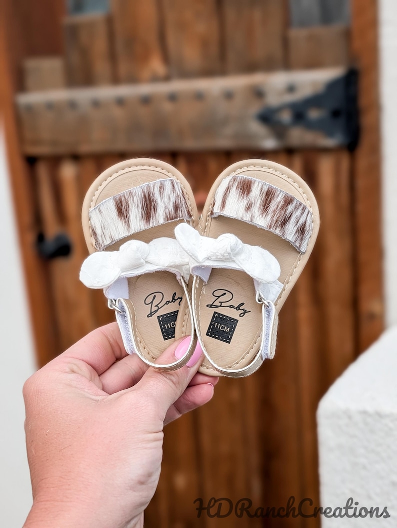 May include: A pair of baby sandals with a tan leather sole and a white strap. The sandals feature a brown and white cowhide design on the top strap and a white bow detail. The soles are marked "Baby" and "11CM".