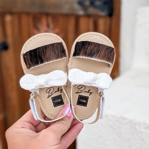 May include: A pair of baby sandals with tan leather soles and a strip of brown and black fur across the top. Each sandal has a white bow and a strap. The word "Baby" and the measurement "11CM" are printed on the sole.