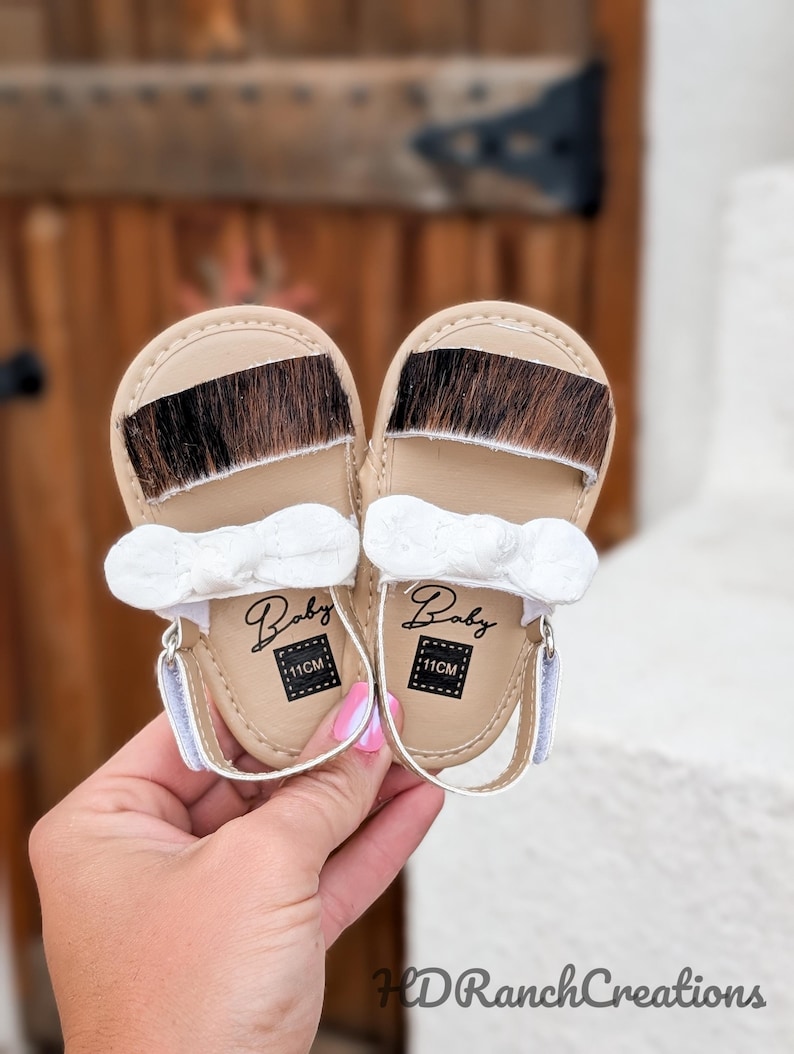 May include: A pair of baby sandals with tan leather soles and a strip of brown and black fur across the top. Each sandal has a white bow and a strap. The word "Baby" and the measurement "11CM" are printed on the sole.