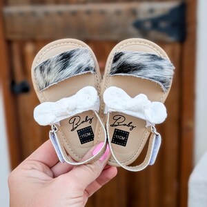 May include: A pair of baby sandals with a tan leather base, featuring a black and white fur accent across the top. Each sandal has a white bow and the word "Baby" printed on the insole, along with a "11CM" label. The sandals are held by a person.