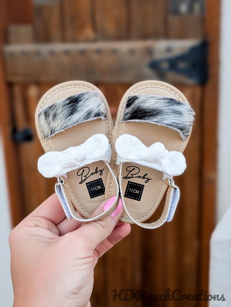 May include: A pair of baby sandals with a tan leather base, featuring a black and white fur accent across the top. Each sandal has a white bow and the word "Baby" printed on the insole, along with a "11CM" label. The sandals are held by a person.