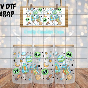 May include: Three glass beverage cups with lids and straws, featuring a colorful space-themed design. The design includes aliens, planets, rockets, stars, and moons. A matching design is displayed on a small wooden sign. The text "UV DTF WRAP" is visible.