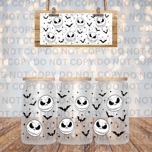 May include: Three glass tumblers with bamboo lids and straws, decorated with a black and white Jack Skellington face and bat pattern. A matching sign hangs above. The background is a blurred bokeh effect.