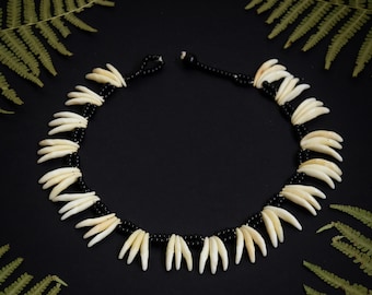 May include: A necklace made of white teeth and black beads. The necklace is laid out on a black background with green fern leaves on either side.