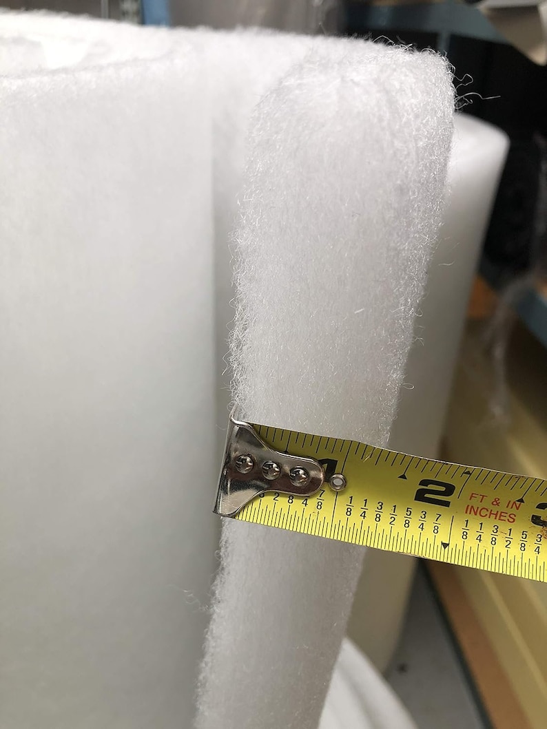 May include: A white, fluffy, rectangular piece of batting material is measured with a yellow tape measure. The tape measure reads 2 feet and 7 inches.
