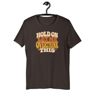 Pode incluir: Uma t-shirt de manga curta castanho-escura com o texto "HOLD ON LET ME OVERTHINK THIS" impresso numa fonte empilhada de estilo retro. As letras utilizam uma paleta de cores creme, laranja e amarelo, centrada no peito da peça de vestuário.