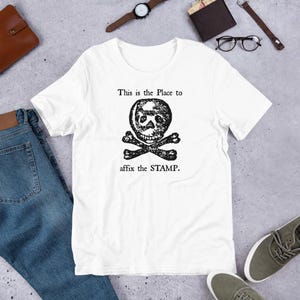 May include: White t-shirt with a black and white skull and crossbones graphic. The text "This is the Place to affix the STAMP." is printed below the graphic.