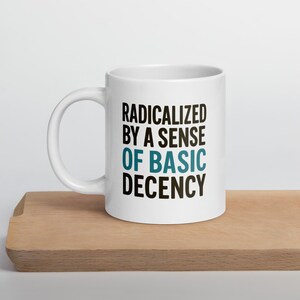 May include: White ceramic mug with the text "RADICALIZED BY A SENSE OF BASIC DECENCY" in black and teal lettering. The mug is on a wooden surface. The mug has a curved handle.