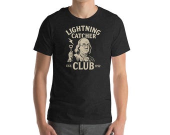 May include: A dark heather grey t-shirt with a graphic of Benjamin Franklin holding a key and lightning bolt. The text "Lightning Catcher Club Est. 1752" is printed on the shirt.