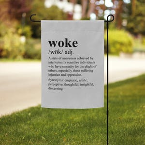 May include: A white garden flag with black text defining the word "woke" as a state of awareness achieved by intellectually sensitive individuals who have empathy for the plight of others, especially those suffering injustice and oppression. Synonyms include: emphatic, astute, perceptive, thoughtful, insightful, and discerning.