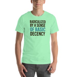 May include: A light green t-shirt with the text "RADICALIZED BY A SENSE OF BASIC DECENCY" in black and blue. The shirt is short-sleeved and appears to be made of a soft material.