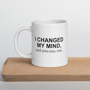 May include: White ceramic coffee mug with the text "I CHANGED MY MIND, and you can, too." in black. The mug is on a wooden surface.