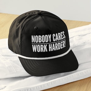 May include: Black baseball cap with a white rope trim and white embroidered text that reads "Nobody Cares. Work Harder!"