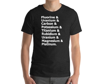 May include: Dark gray t-shirt with white text listing chemical elements: Fluorine, Uranium, Carbon, Potassium, Titanium, Rubidium, Uranium, Magnesium, and Platinum. The shirt is short-sleeved and has a crew neck.
