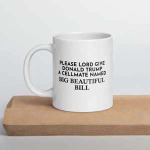May include: White ceramic mug with the text "PLEASE LORD GIVE DONALD TRUMP A CELLMATE NAMED BIG BEAUTIFUL BILL" in black, set on a wooden surface.