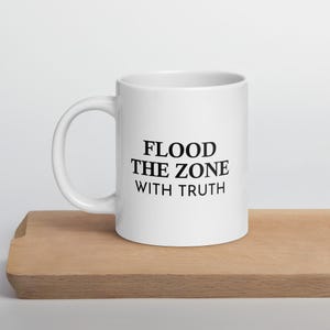 May include: White ceramic coffee mug with a black inscription that reads "Flood the Zone with Truth".