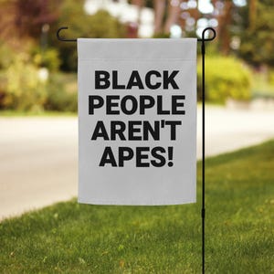 Peut inclure: Un drapeau de jardin gris clair avec le texte "BLACK PEOPLE AREN'T APES!" en lettres noires en gras. Le drapeau est suspendu à un piquet de jardin en métal noir, sur fond d'herbe verte et d'arrière-plan flou.