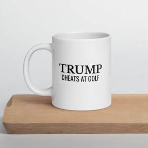 May include: White ceramic coffee mug with a black inscription that reads "TRUMP CHEATS AT GOLF".