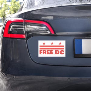 May include: A rectangular white sticker with red text and design, affixed to the rear of a dark gray car. The sticker reads "FREE DC" below three red stars and horizontal lines.