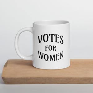 May include: White ceramic mug with the words "VOTES FOR WOMEN" in black, displayed on a wooden surface. The mug has a curved handle and a cylindrical shape. The text is in a bold, stylized font.