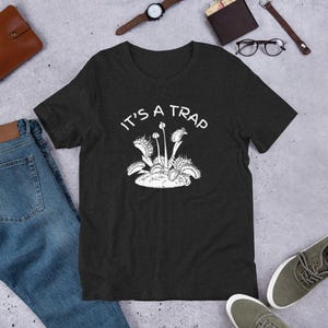 May include: A dark grey t-shirt with a white graphic of a Venus flytrap and the text "It's a Trap".