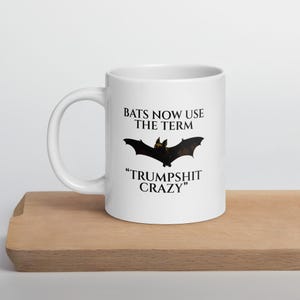 May include: White ceramic mug with a black bat graphic and the text "BATS NOW USE THE TERM" above the bat and "TRUMPSHIT CRAZY" below. The mug is on a wooden surface.