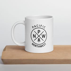 May include: A white ceramic mug with a black graphic that reads "PACIFIC NORTHWEST." The design includes the letters "P," "N," and "W" within a circle, crossed by lines, and a small tree icon. The mug is on a wooden surface.