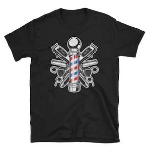 Puede incluir: Camiseta negra con una ilustraci&oacute;n vintage de un poste de barbero con herramientas de barbero cruzadas. El poste de barbero es rojo, blanco y azul con una base plateada. Las herramientas incluyen un par de tijeras, una navaja de afeitar y dos cortadoras.