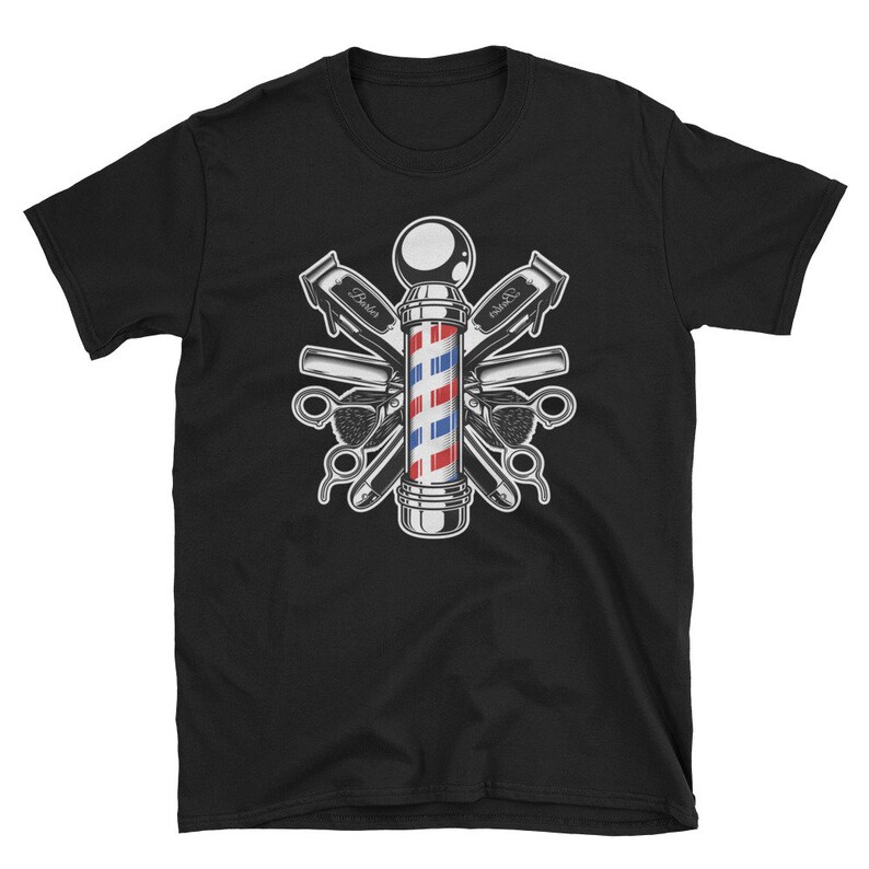 Puede incluir: Camiseta negra con una ilustraci&oacute;n vintage de un poste de barbero con herramientas de barbero cruzadas. El poste de barbero es rojo, blanco y azul con una base plateada. Las herramientas incluyen un par de tijeras, una navaja de afeitar y dos cortadoras.