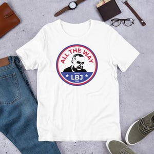 May include: White t-shirt with a blue and red circular graphic featuring a portrait of Lyndon B. Johnson and the text "All the Way with LBJ 1964".